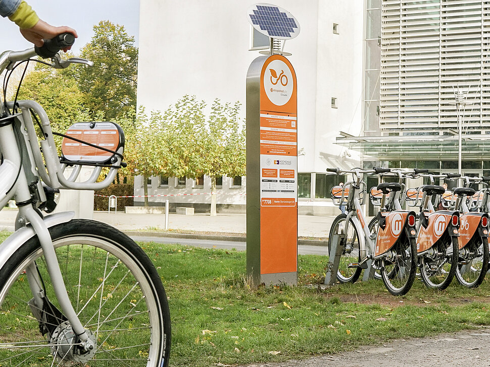 Leihstation mit vielen Fahrrädern vor einem großen Gebäude in urbaner Umgebung. 