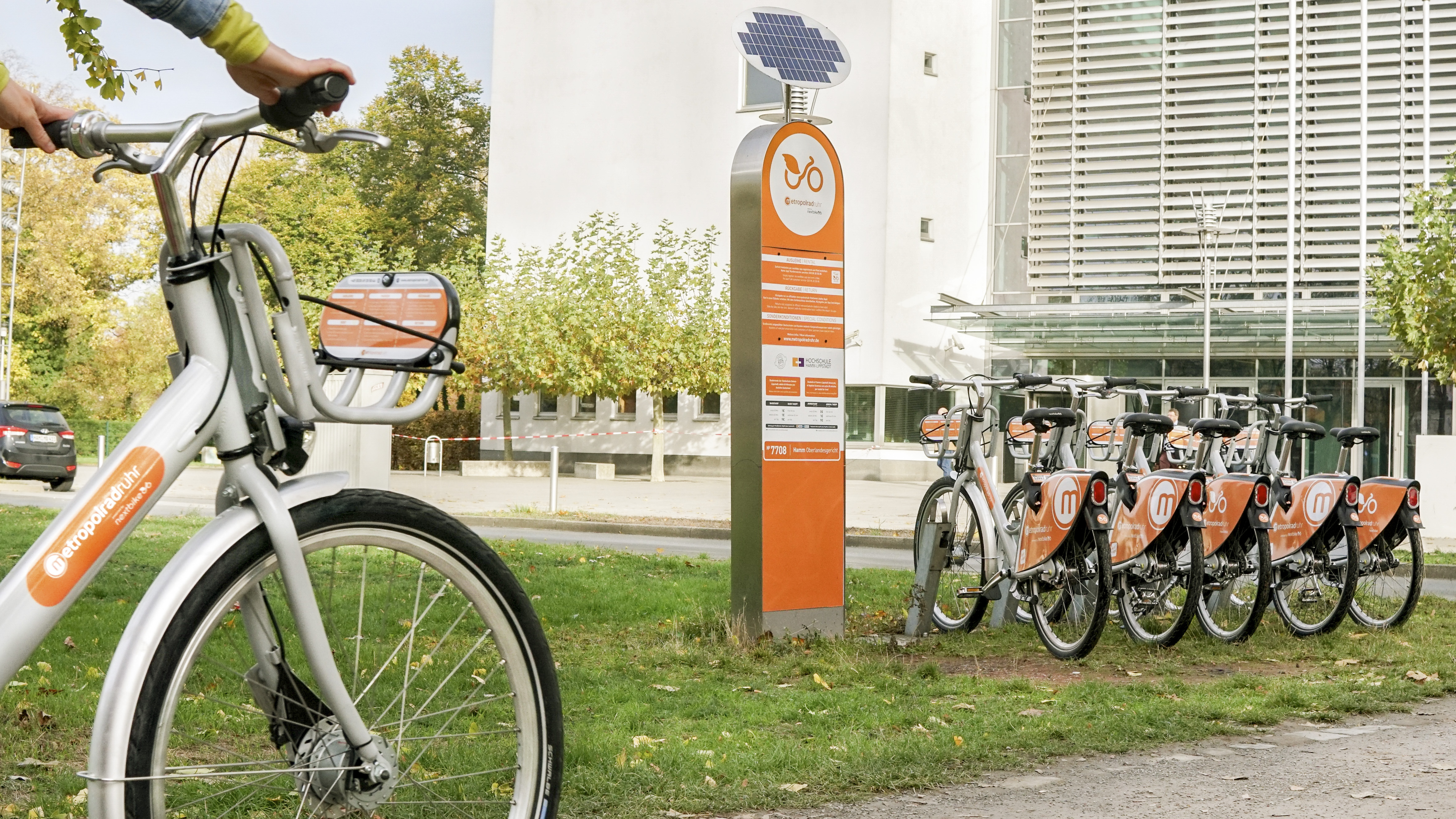 Leihstation mit vielen Fahrrädern vor einem großen Gebäude in urbaner Umgebung. 